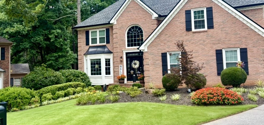 Front-yard landscape installation with layered foundation beds, shaped shrubs, flowering plants and a brick home